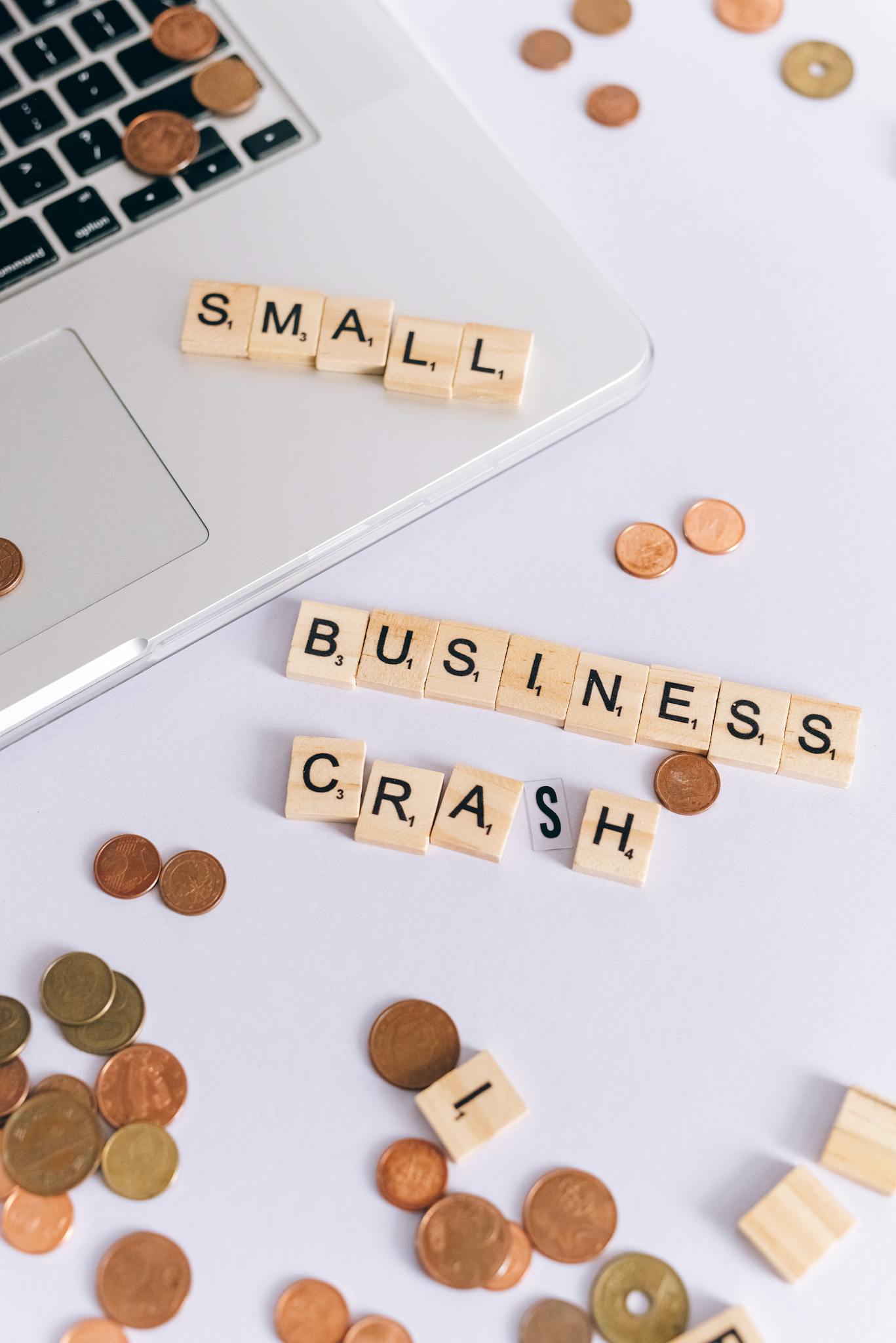 Scrabble tiles spelling 'Small Business Crash' with coins on a laptop, symbolizing financial struggle.